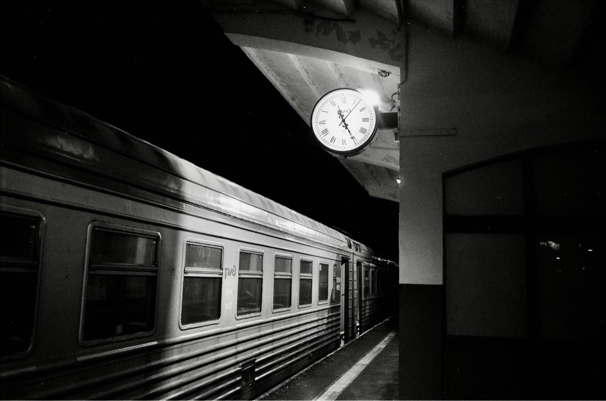 Black-and-white photograph of a train at night beneath an illuminated station clock