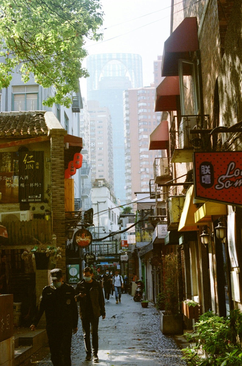 Sunlit city alley with pedestrians, shop signs, and tall buildings in the distance