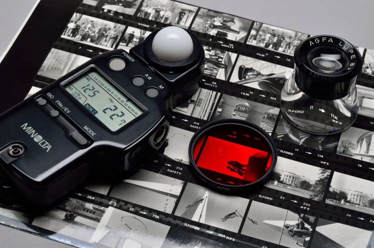 Handheld Minolta light meter resting on black-and-white contact sheets beside a magnifying loupe