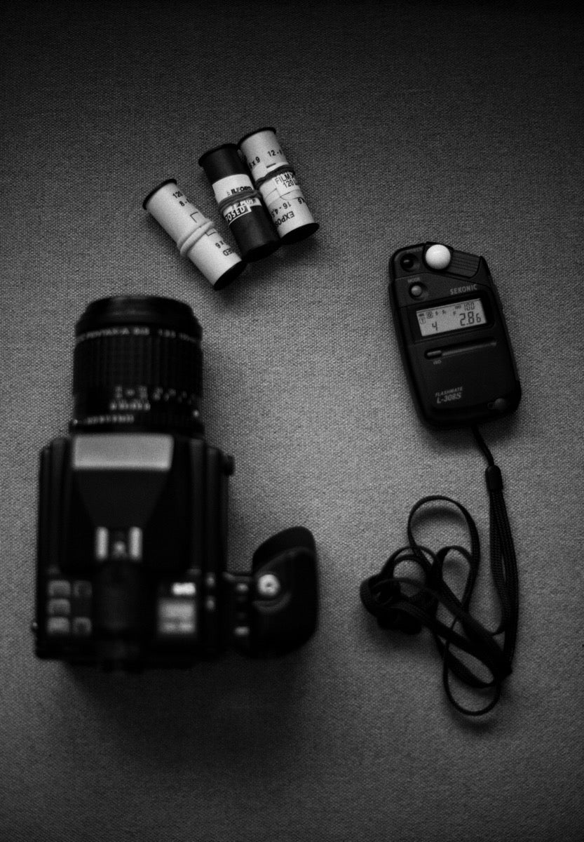 Black-and-white photograph of a camera, rolls of film, and a handheld light meter arranged on fabric
